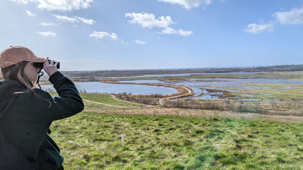 Woman with cap on looks through binoculars. A view overlooking RSPB St Aidan's is visible in the background.