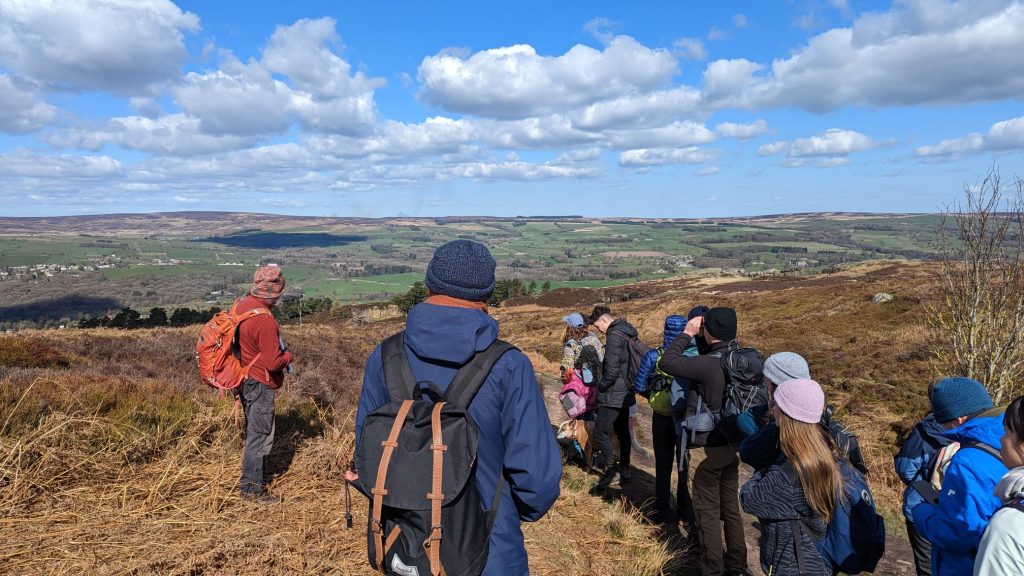 A group of people stand atop a hill, looking off into the distance where green fields and hills can be seen.